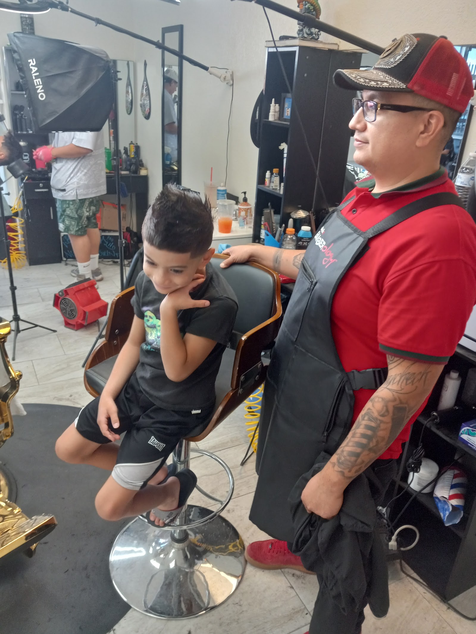Barber in a red shirt and apron standing beside a young boy with a spiky haircut in the chair